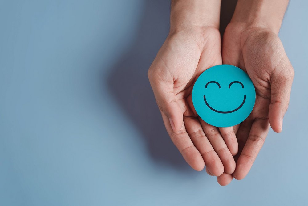 Ein lachender Smiley auf blauem Papier wird von zwei Händen präsentiert.