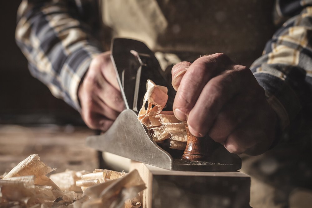 Zu sehen sind zwei Männerhände die mit einem alten Hobel ein großes Stück Holz bearbeiten.