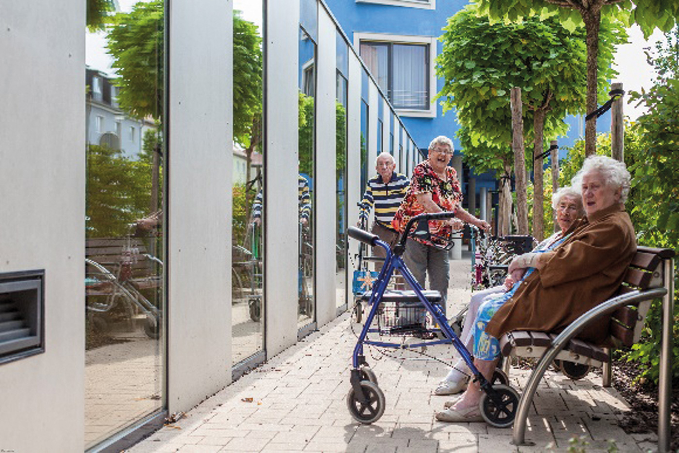 Seniorinnen sitzen auf einer Bank vor einem Gebäude, vor ihnen steht ihre Gehiilfe. Im Hintergrund laufen eine ältere Frau und ein Senior. Alle Blicken in die Kamera.