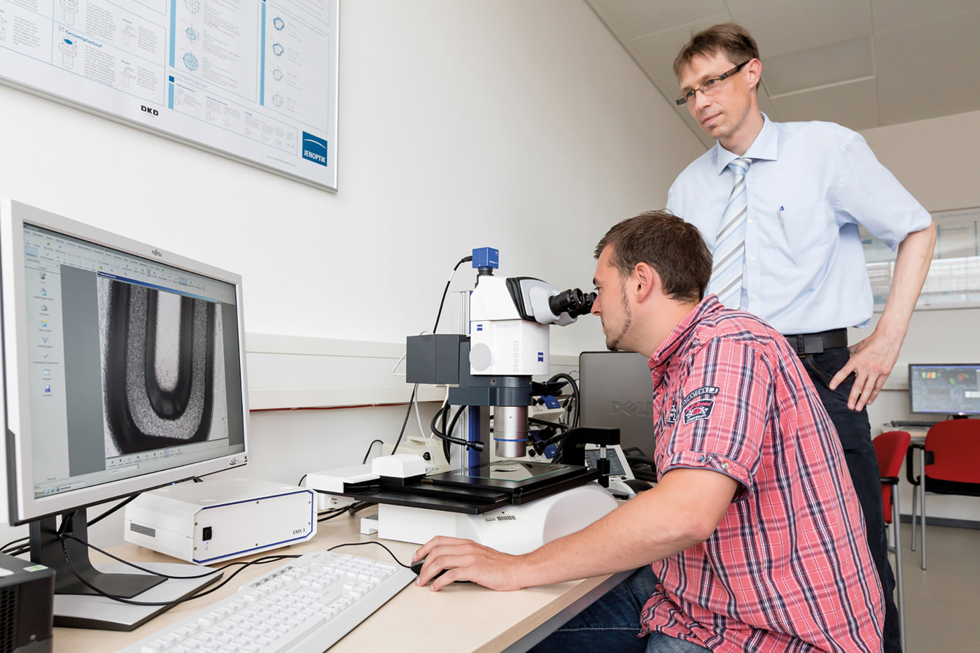 Ein Mann guckt in ein Miskroskop, links von ihm steht ein Bildschirm auf dem das Mikroskopbild zu sehen ist,hinter ihm steht ein Mann und guckt auf den Bildschirm